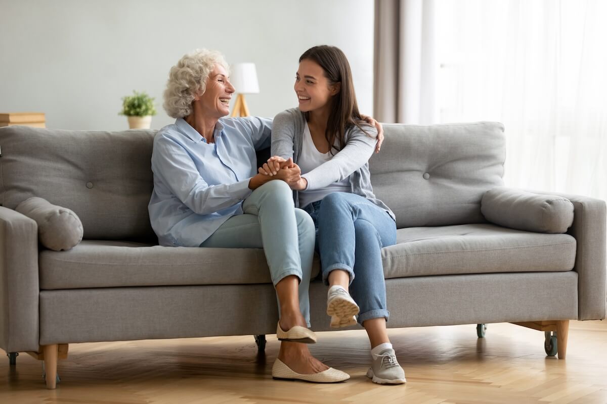 What if Mom Doesn’t Want to Move to Senior Living adult granddaughter and senior grandmother having fun enjoying talk sit on sofa in modern living room, smiling old mother hugging young grown daughter bonding chatting relaxing at home together