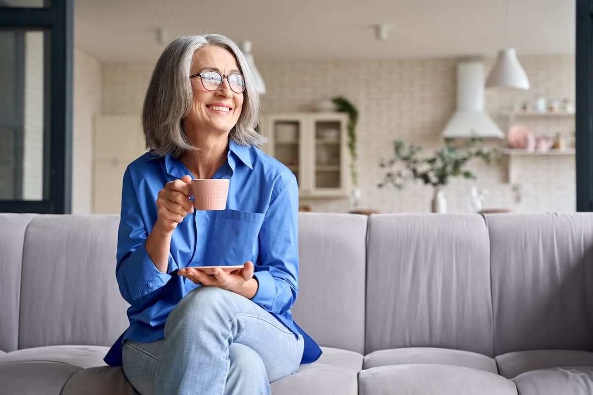 stress management tips Smiling mature elder 60s woman sitting relaxing with cup of tea, coffee. Senior mid age stylish look woman with eyeglasses portrait with cup looking away at modern home.
