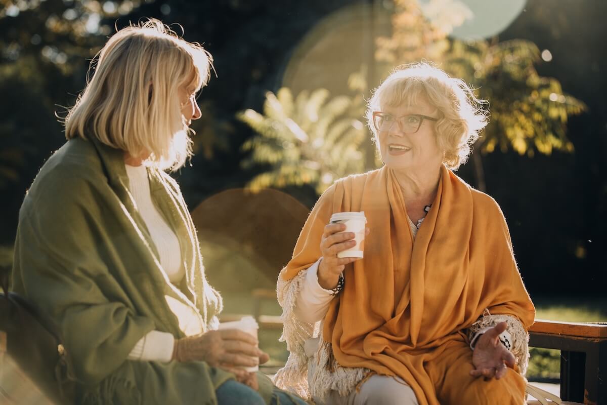Two senior women are sharing a moment of friendship, sitting on a bench in a sunny autumn park, sipping coffee and engaging in lively conversation senior living community winter